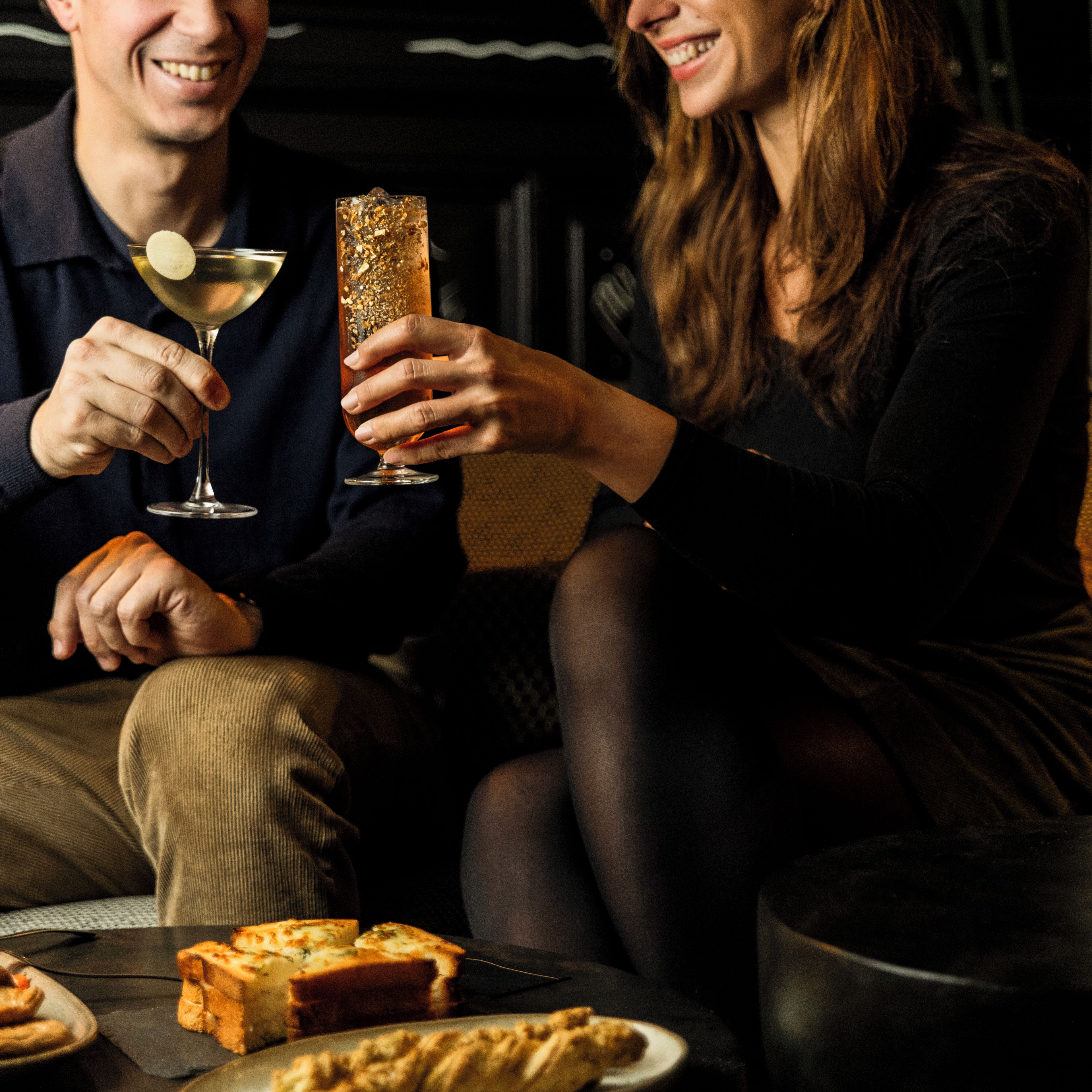 A couple sitting on a couch with a glass of wine and a bottle of champagne. There are slices of pizza on a plate in front of them.