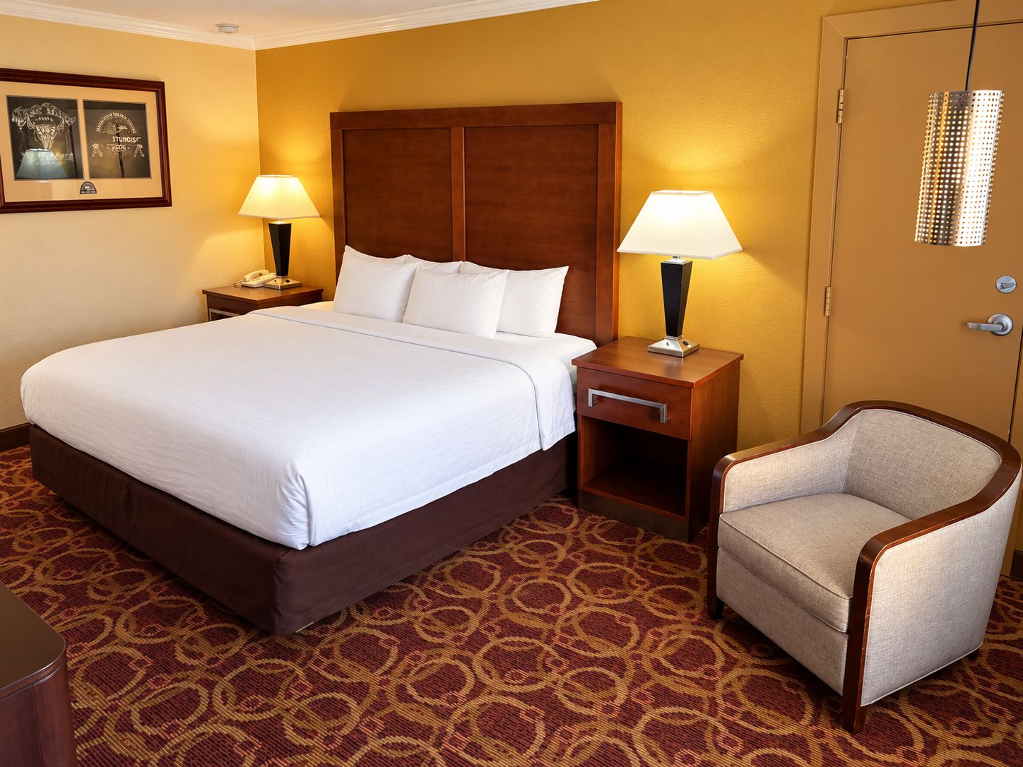 Una habitación de hotel con una cama grande, dos mesitas de noche con lámparas, una silla y suelo alfombrado.