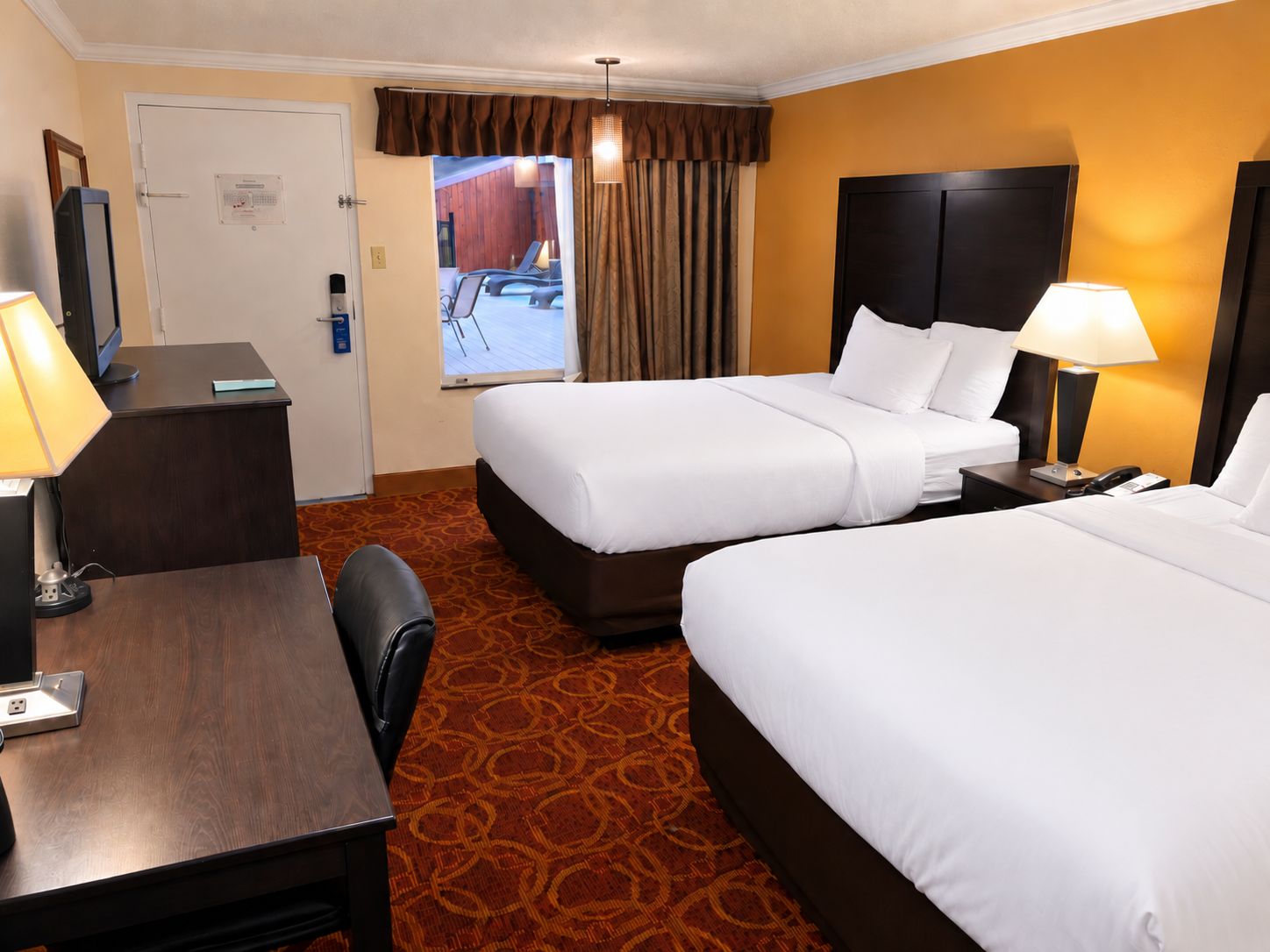 A hotel room with two beds, a desk, a chair, and a view of an outdoor area.