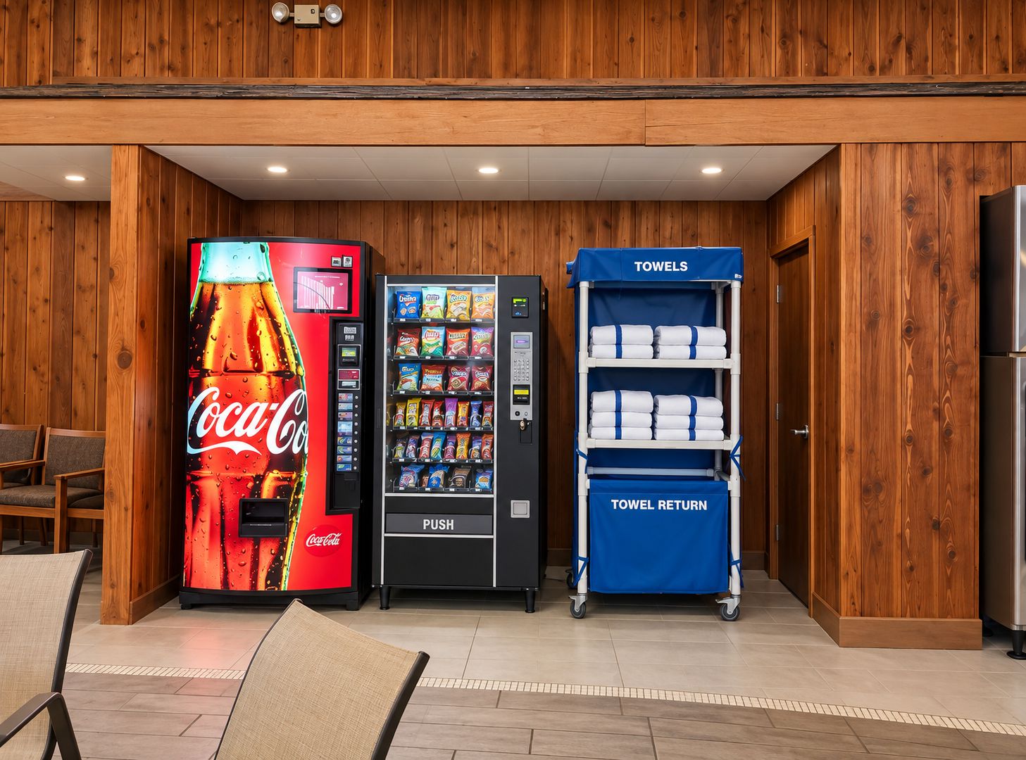 A vending area with a Coca-Cola machine, a snack vending machine, and a towel return cart in a wooden-finished room.