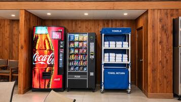 A vending area with a Coca-Cola machine, a snack vending machine, and a towel return cart in a wooden-finished room.