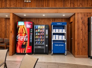 A vending area with a Coca-Cola machine, a snack vending machine, and a towel return cart in a wooden-finished room. - Sturgis Lodge & Suites, Travelodge by Wyndham (Sturgis)