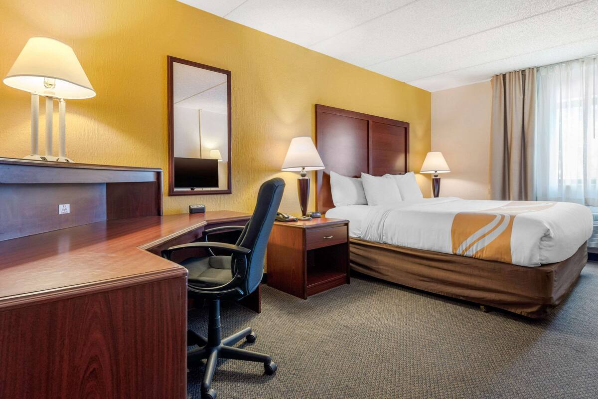 A hotel room with a wooden desk, two lamps, a bed with white and brown bedding, a chair, and a large mirror on the wall.
