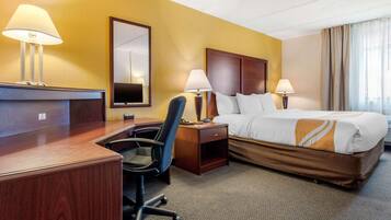 Habitación de hotel con un escritorio de madera, dos lámparas, una cama con ropa de cama blanca y marrón, una silla y un espejo grande en la pared.