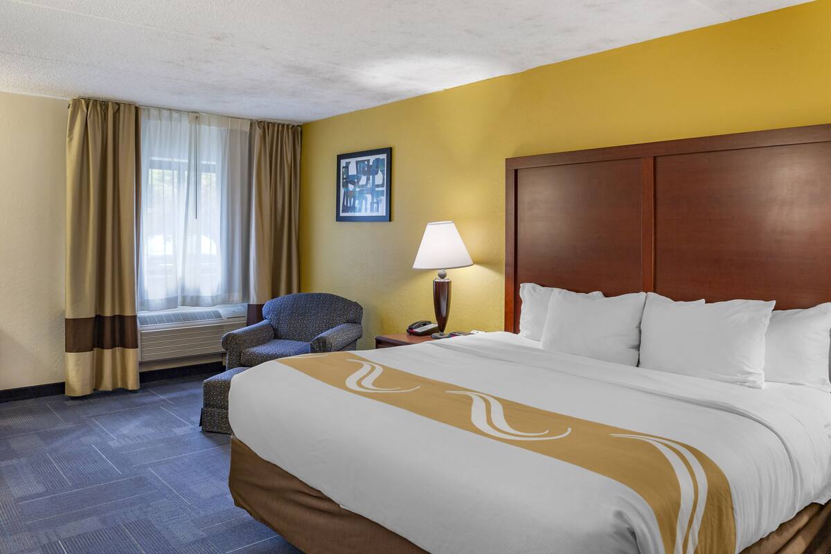 A hotel room with a large bed, a chair, a nightstand with a lamp, and a window with curtains.