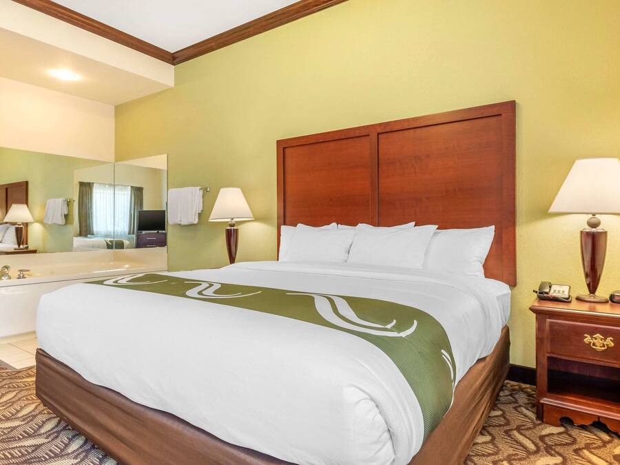 Une chambre d’hôtel avec un grand lit, une tête de lit en bois, deux lampes de chevet, une table de chevet, un miroir et une salle de bain équipée d’un lavabo et de toilettes, ainsi qu’un tapis à motifs.