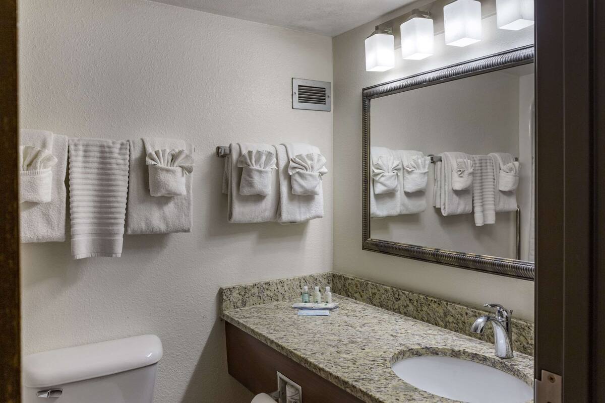 A bathroom with a toilet, sink, and mirror. There are towels hanging on the wall and a vent above the toilet.