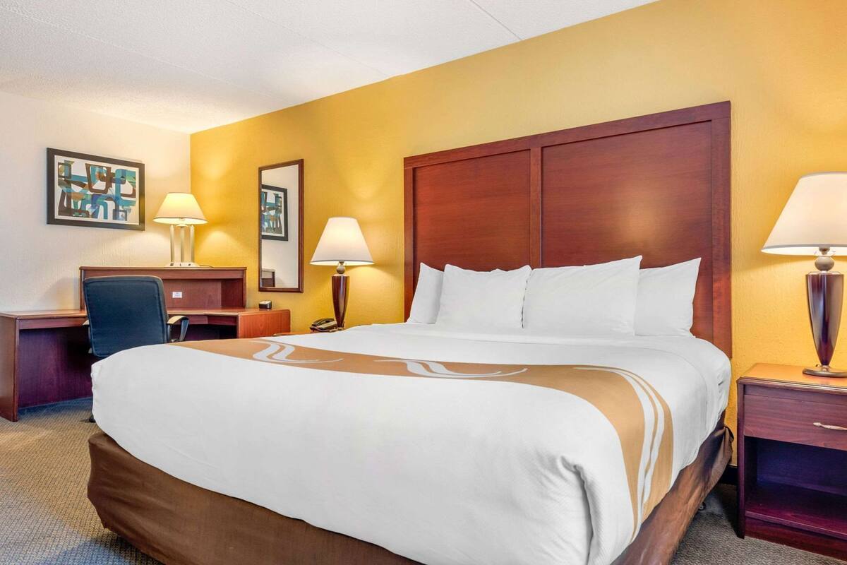 A hotel room with a large bed, two bedside tables with lamps, a desk with a chair, and a wall-mounted picture.