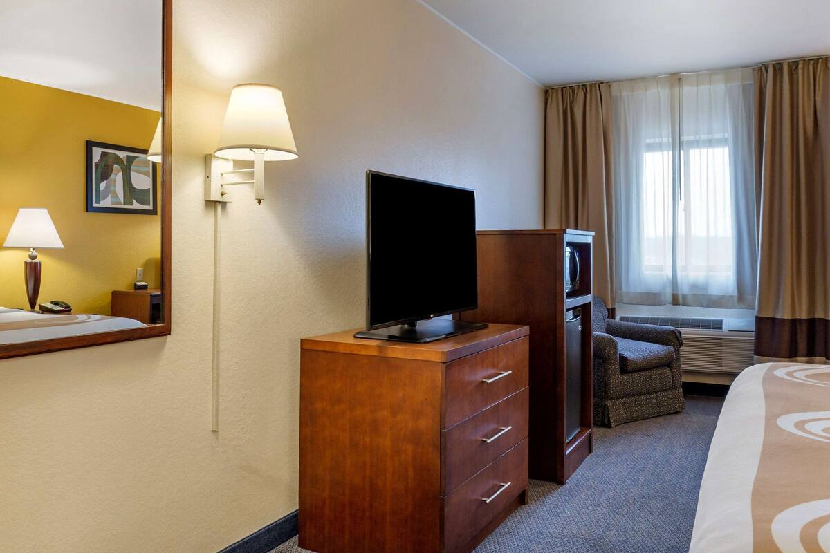 A hotel room with a wooden dresser, a flat-screen TV, a chair, a bed, a desk, a lamp, and a window with curtains.