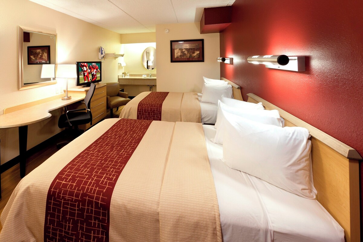 A hotel room with two beds, a desk, a TV, a mirror, and a bathroom.