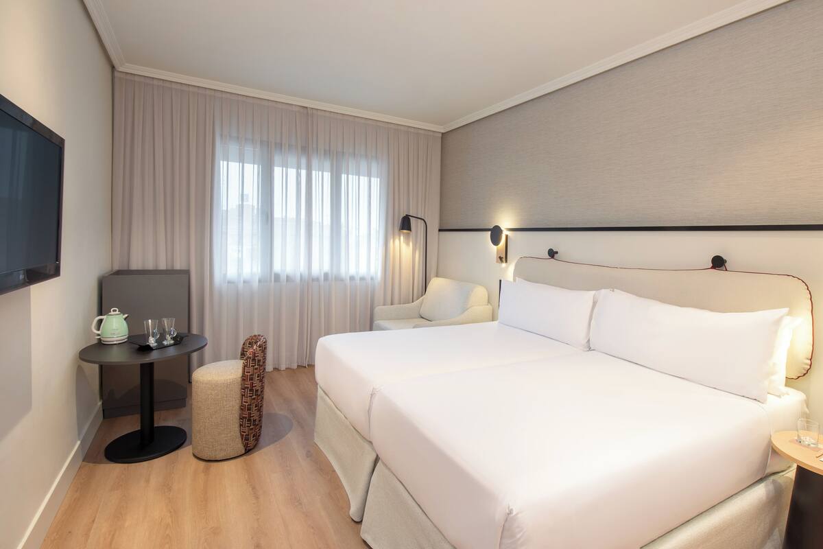 Hotellihuone, jossa on suuri sänky, pieni pöytä, jossa on vedenkeitin, tuoli ja seinälle asennettu televisio.