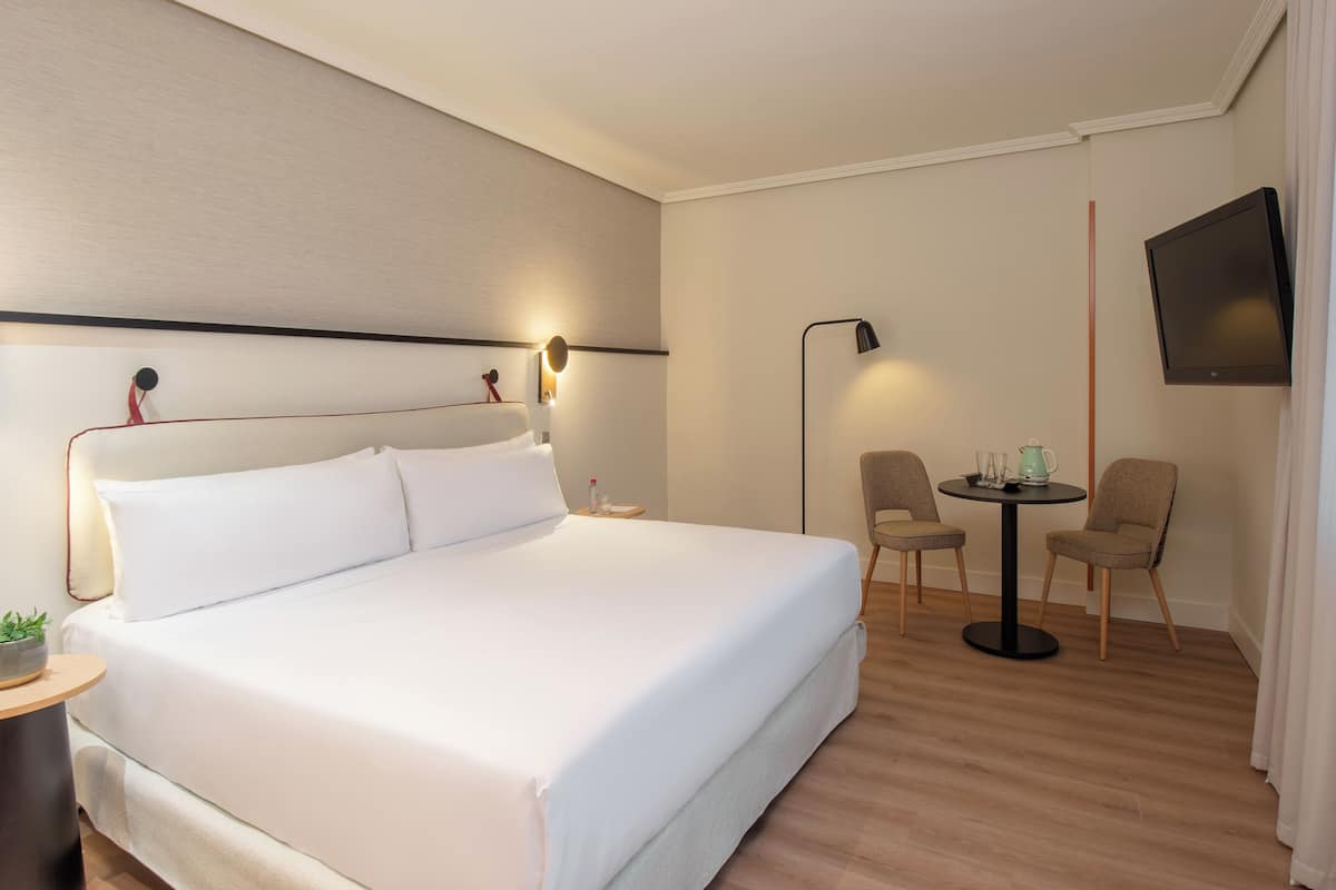 Hotellihuone, jossa on suuri sänky, yöpöytä, lamppu, televisio ja pieni pöytä tuoleineen.