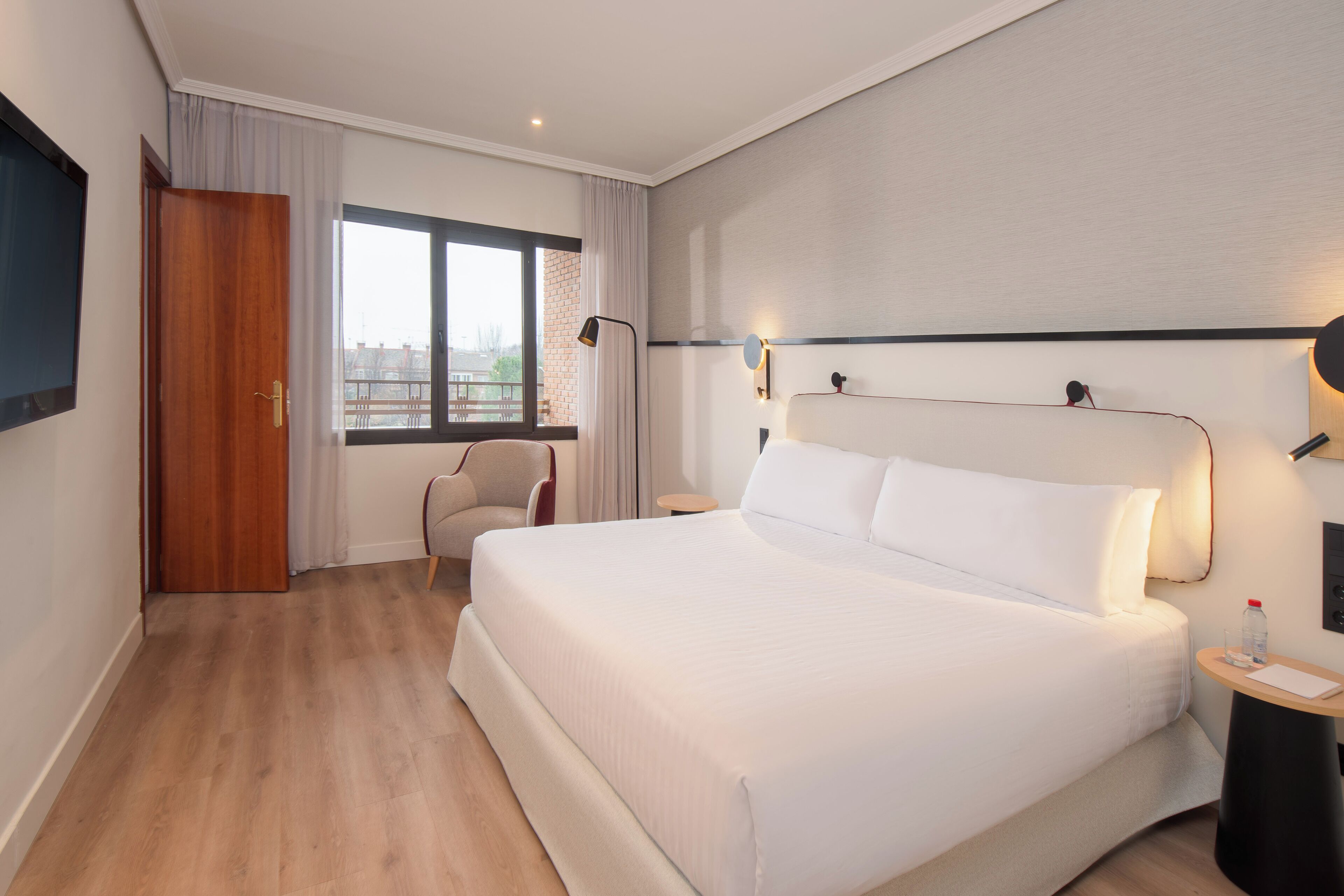 A hotel room with a large bed, a chair, a desk, a window with curtains, and a flat-screen TV mounted on the wall.
