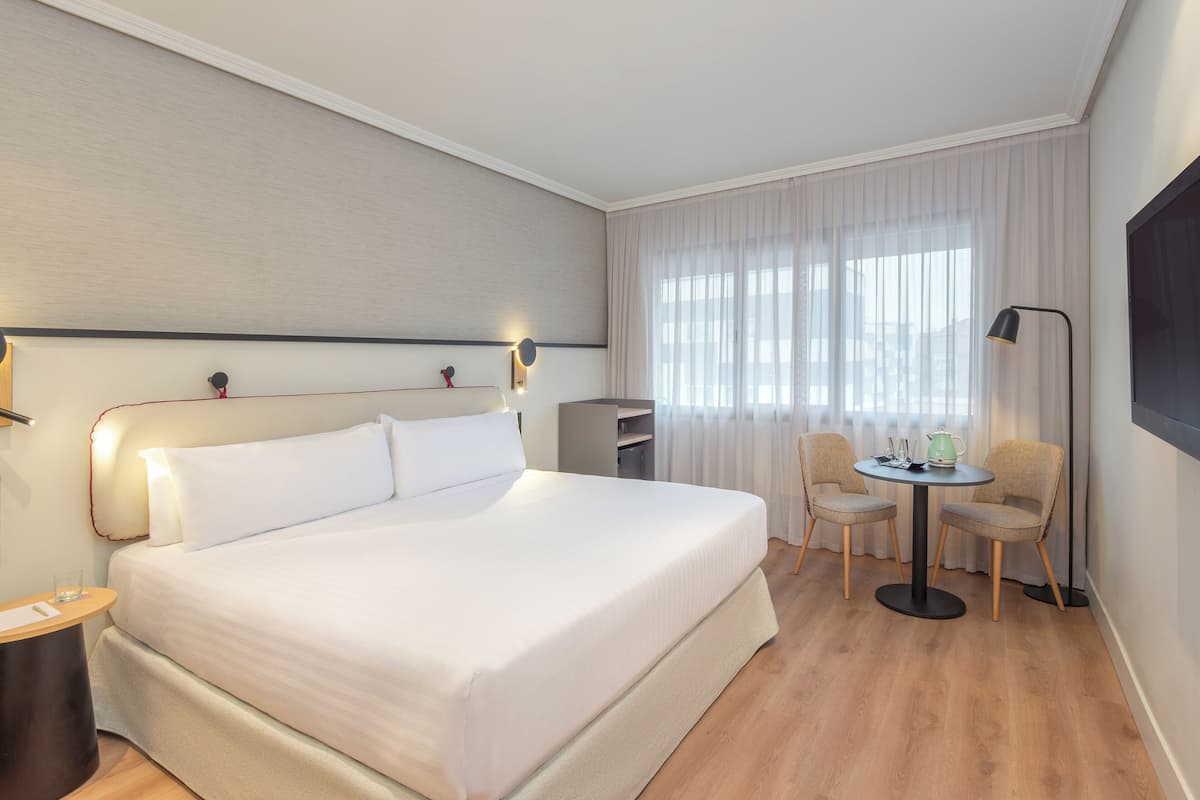 Hotellihuone, jossa on suuri sänky, yöpöytä, pieni pöytä tuoleineen, lamppu ja suuri ikkuna, jossa on verhot.