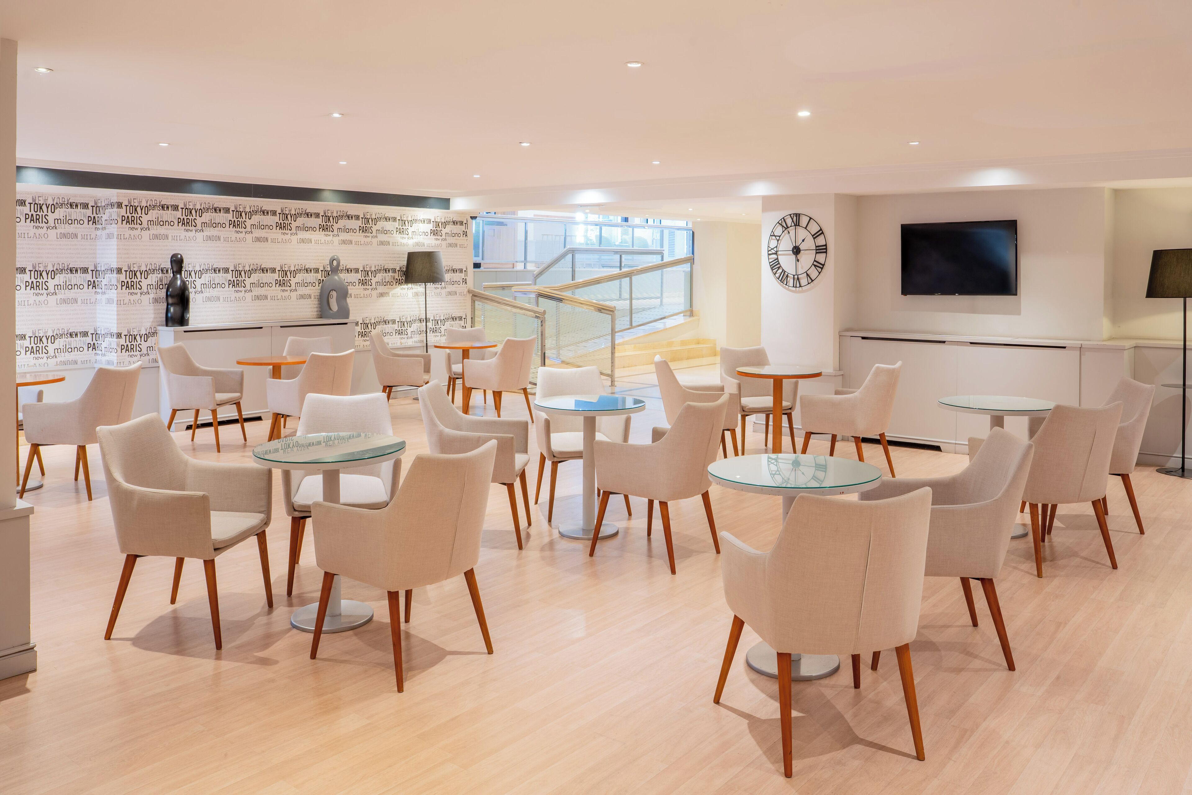 A modern hotel lobby with round tables and chairs, a large clock, and a TV on the wall.
