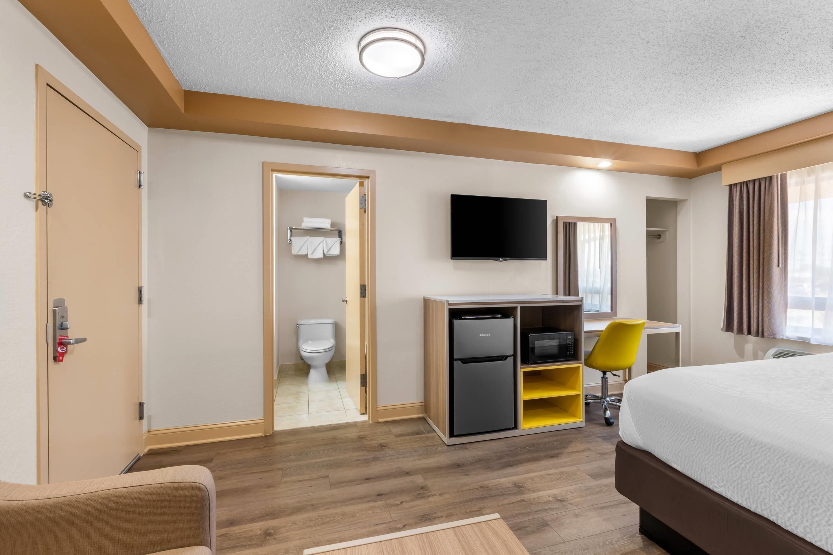 A hotel room with a bed, a desk with a computer, a TV, and a bathroom visible through an open door.
