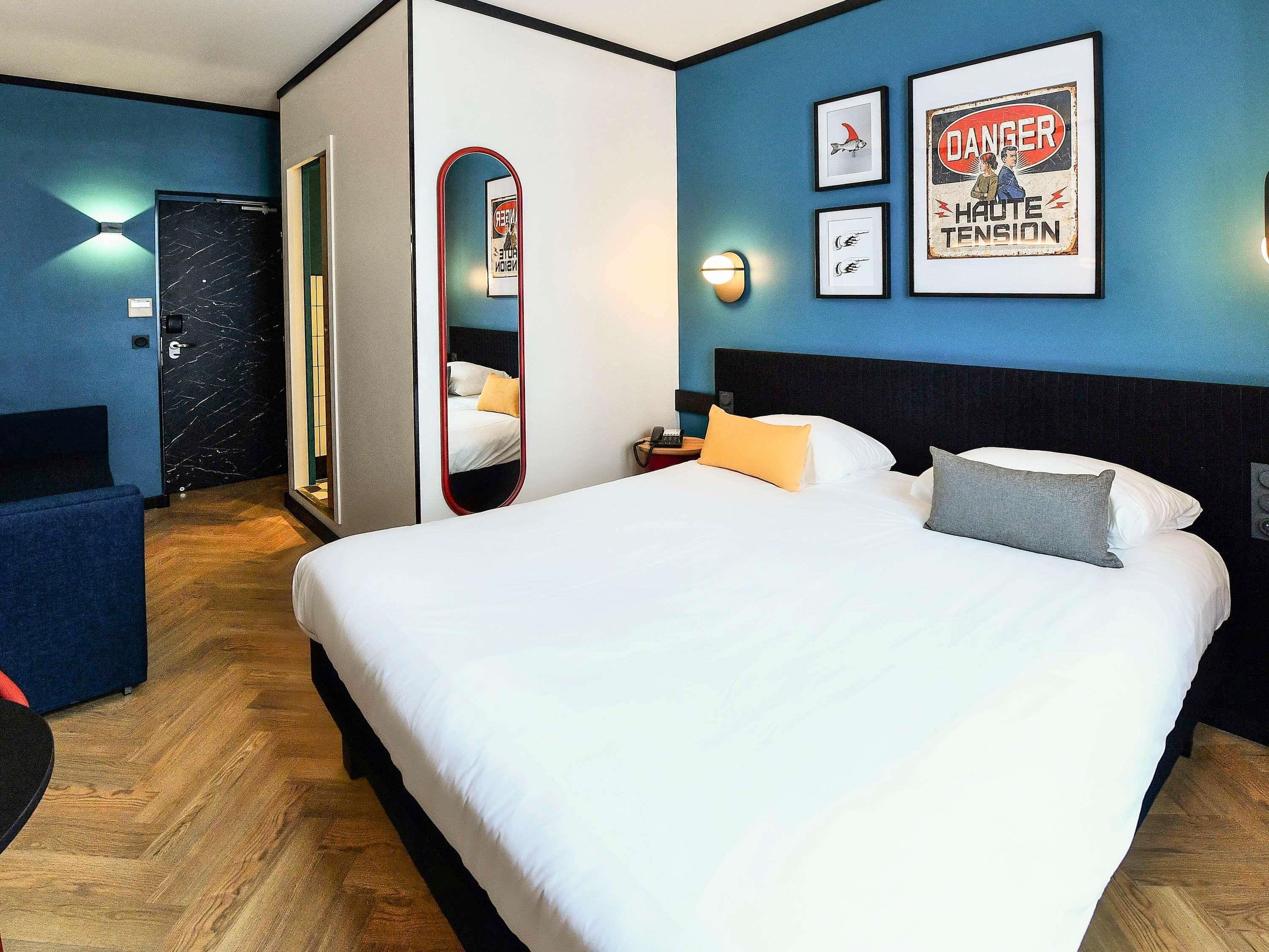 A hotel room with a large bed, a bedside table with a lamp, a mirror, and framed pictures on the wall.