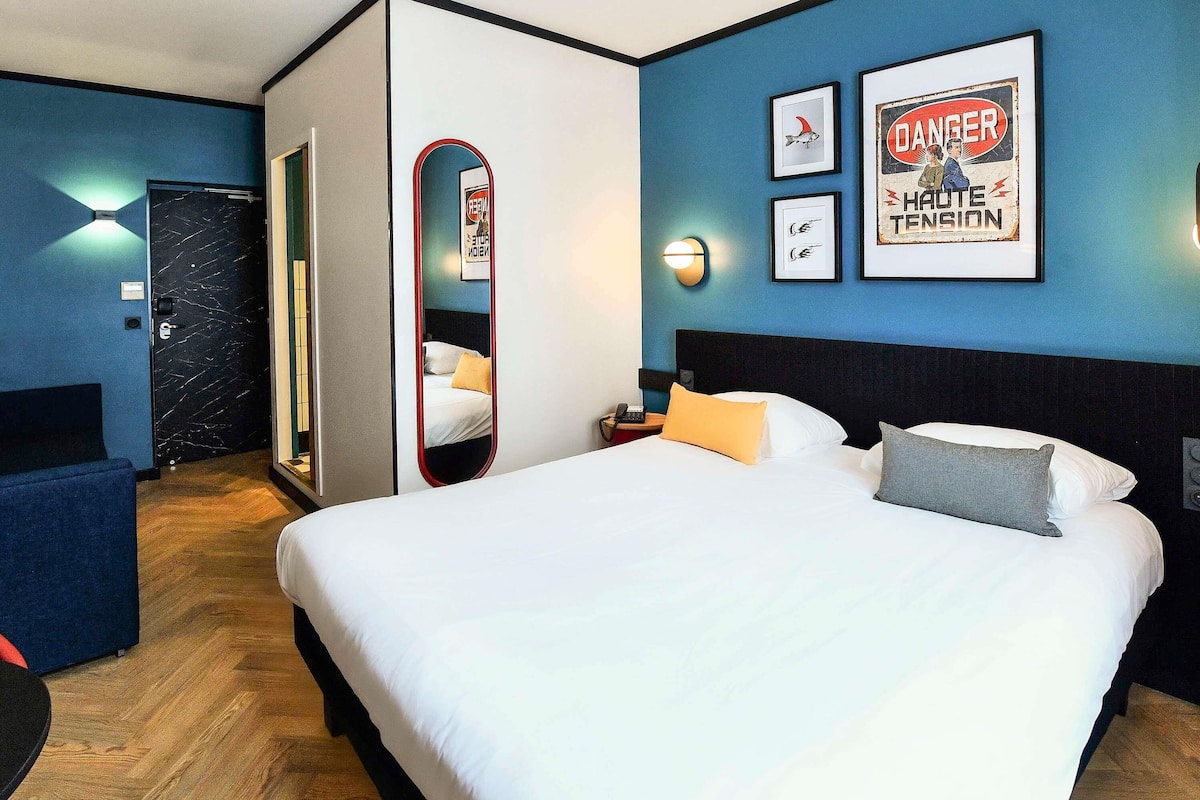 A hotel room with a large bed, a bedside table with a lamp, a mirror, and framed pictures on the wall.