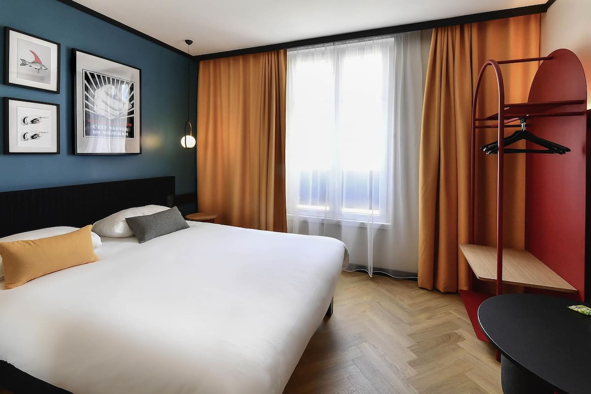 Quarto de hotel com cama, escrivaninha, cadeira, janela com cortinas e decoração nas paredes.