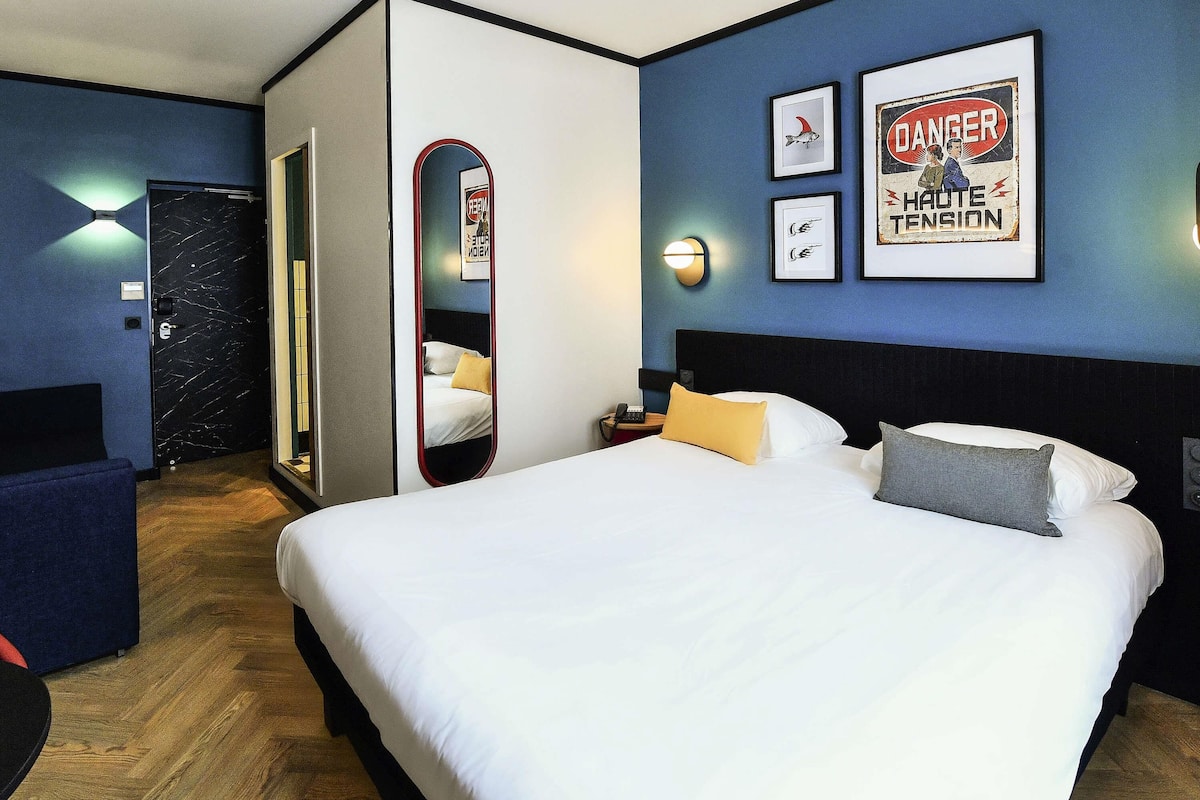 Quarto de hotel com uma cama grande, mesas de cabeceira, um espelho e quadros nas paredes.