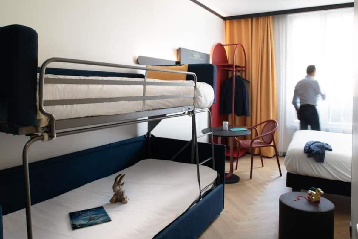 Habitación de hotel con literas, un escritorio, una silla, una mesita y una ventana con cortinas.