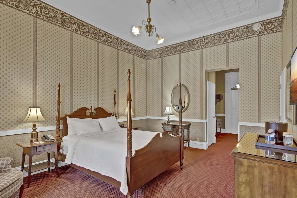 Een hemelbed met houten hoofdbord, nachtkastje met lamp, spiegel en een dressoir met spiegel.