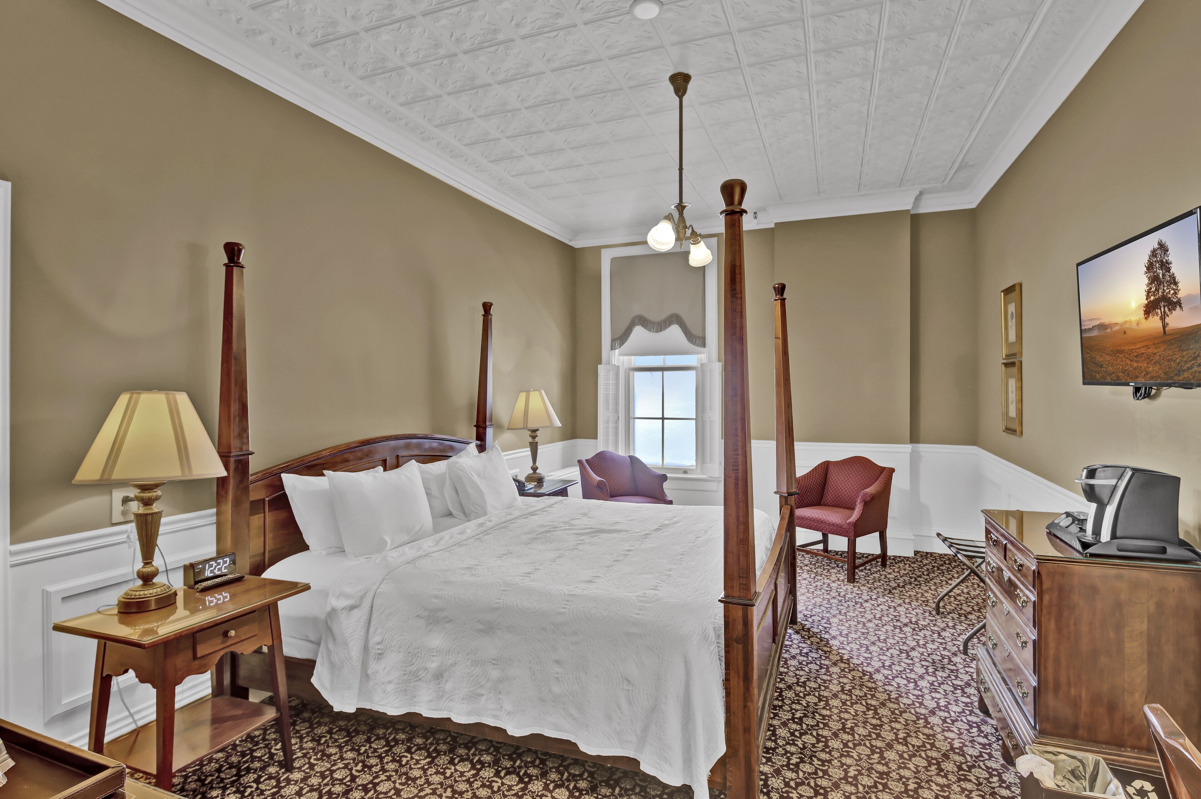 A four-poster bed with a white bedspread, a wooden nightstand with a lamp, a red armchair, a dresser, and a television on the wall.