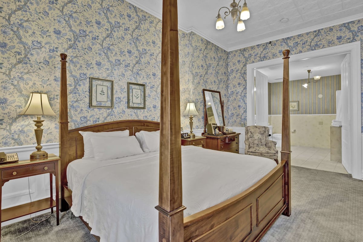 Een kamer met een hemelbed, bloemenbehang, een nachtkastje van hout met een lamp en een spiegel aan de muur.