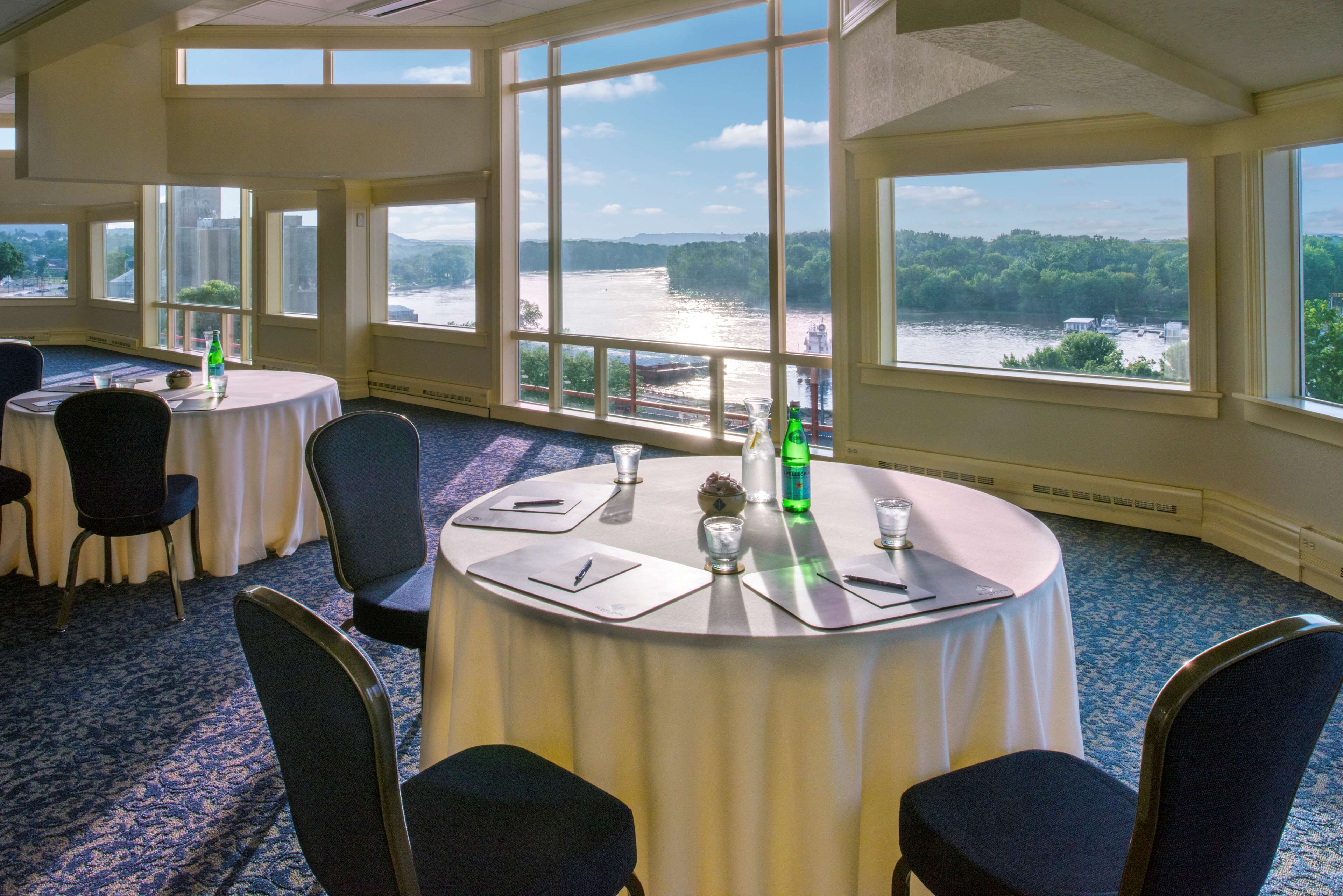 Un salón de conferencias con mesas redondas, sillas y grandes ventanales con vistas al río.