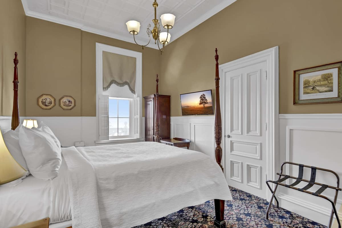 Een slaapkamer met een hemelbed, een kroonluchter, een groot raam en een schilderij aan de muur.