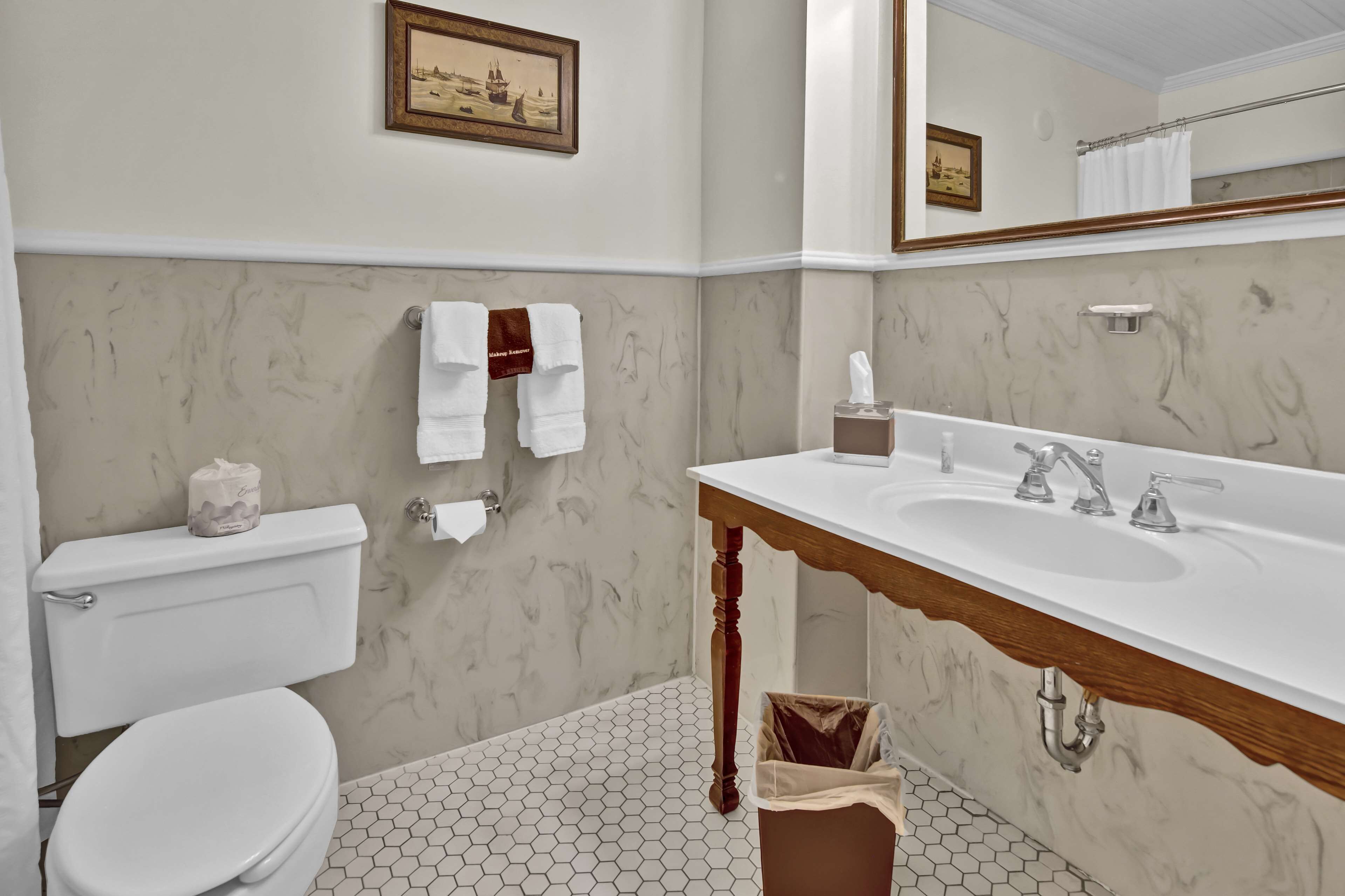 A bathroom with a toilet, sink, and mirror. There are towels hanging on the wall and a picture frame above the mirror.