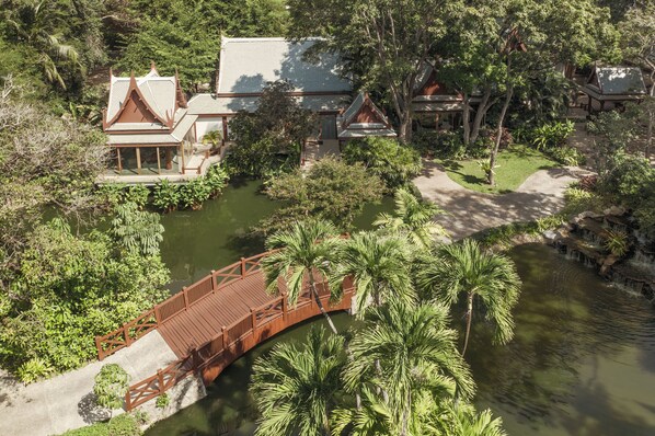 Uma ponte de madeira tradicional sobre um lago, rodeada por vegetação exuberante e edifícios típicos com telhados inclinados.