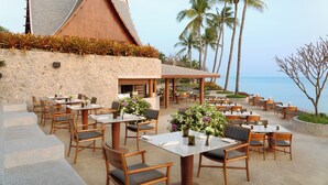 An outdoor dining area with stone walls, wooden tables, and chairs, overlooking a body of water and palm trees.