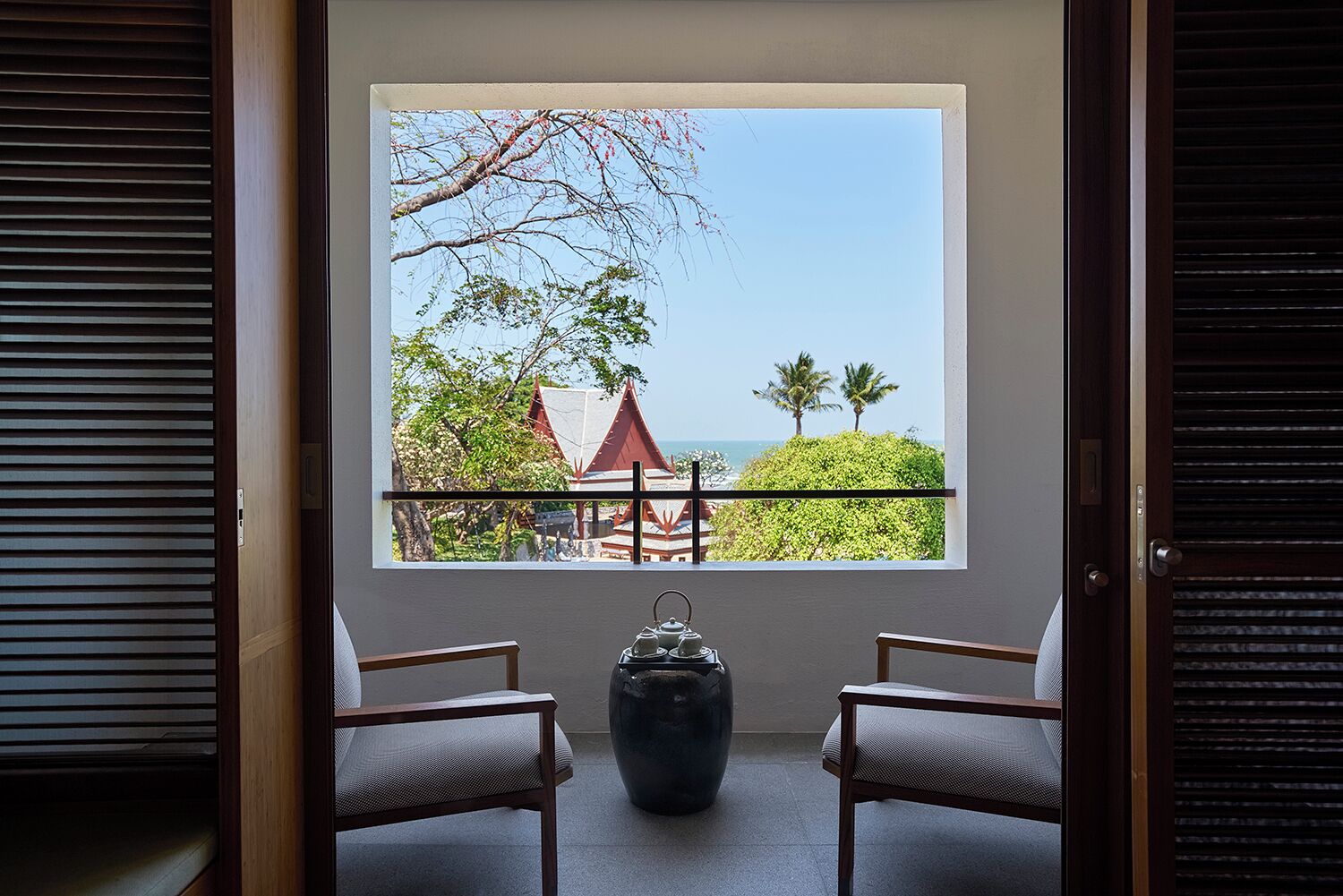 A room with a large window offering a view of a beach, palm trees, and traditional buildings. There are two chairs and a large vase on the floor.