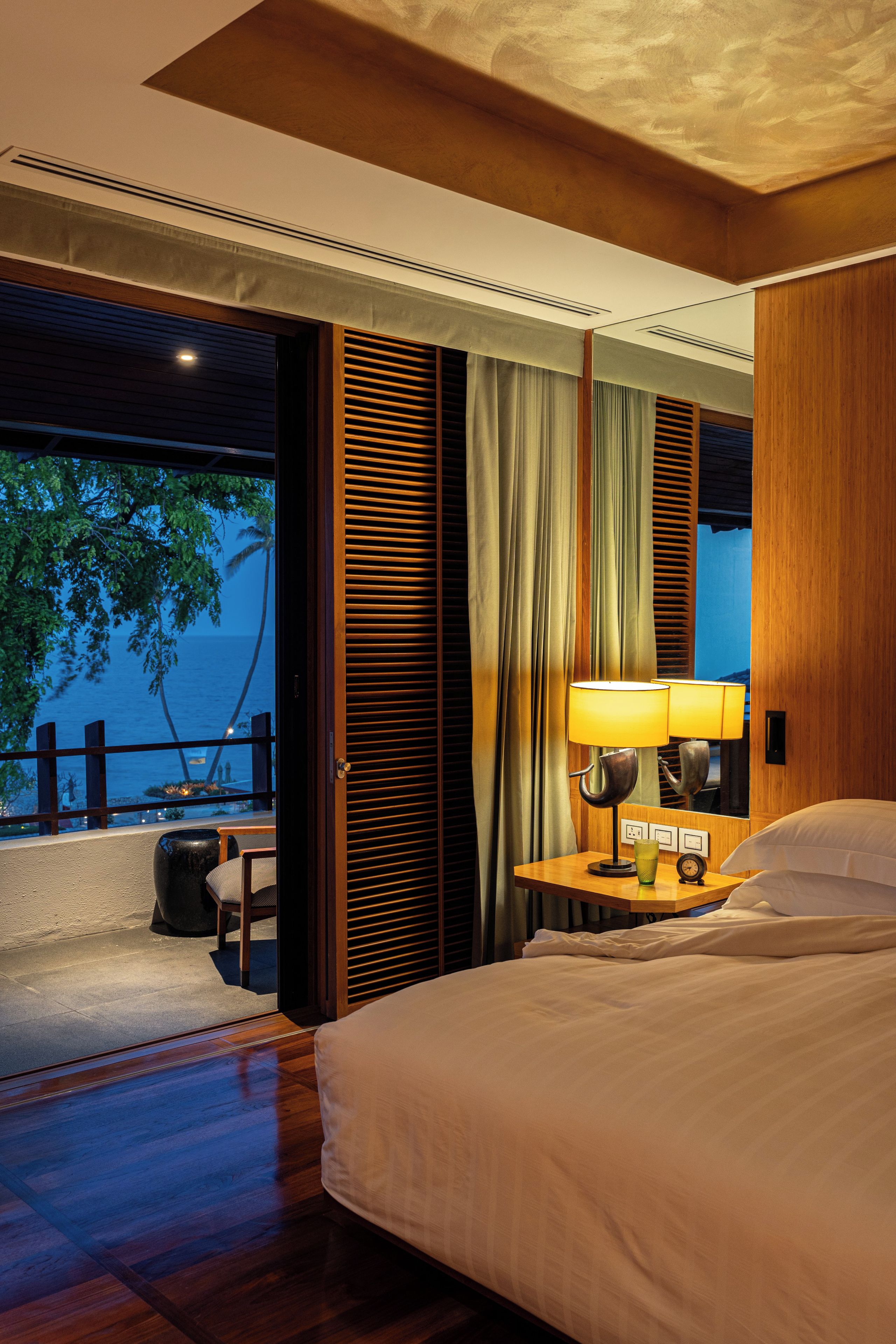 A hotel room with a bed, a nightstand with a lamp, a view of the ocean, and a balcony with a railing.