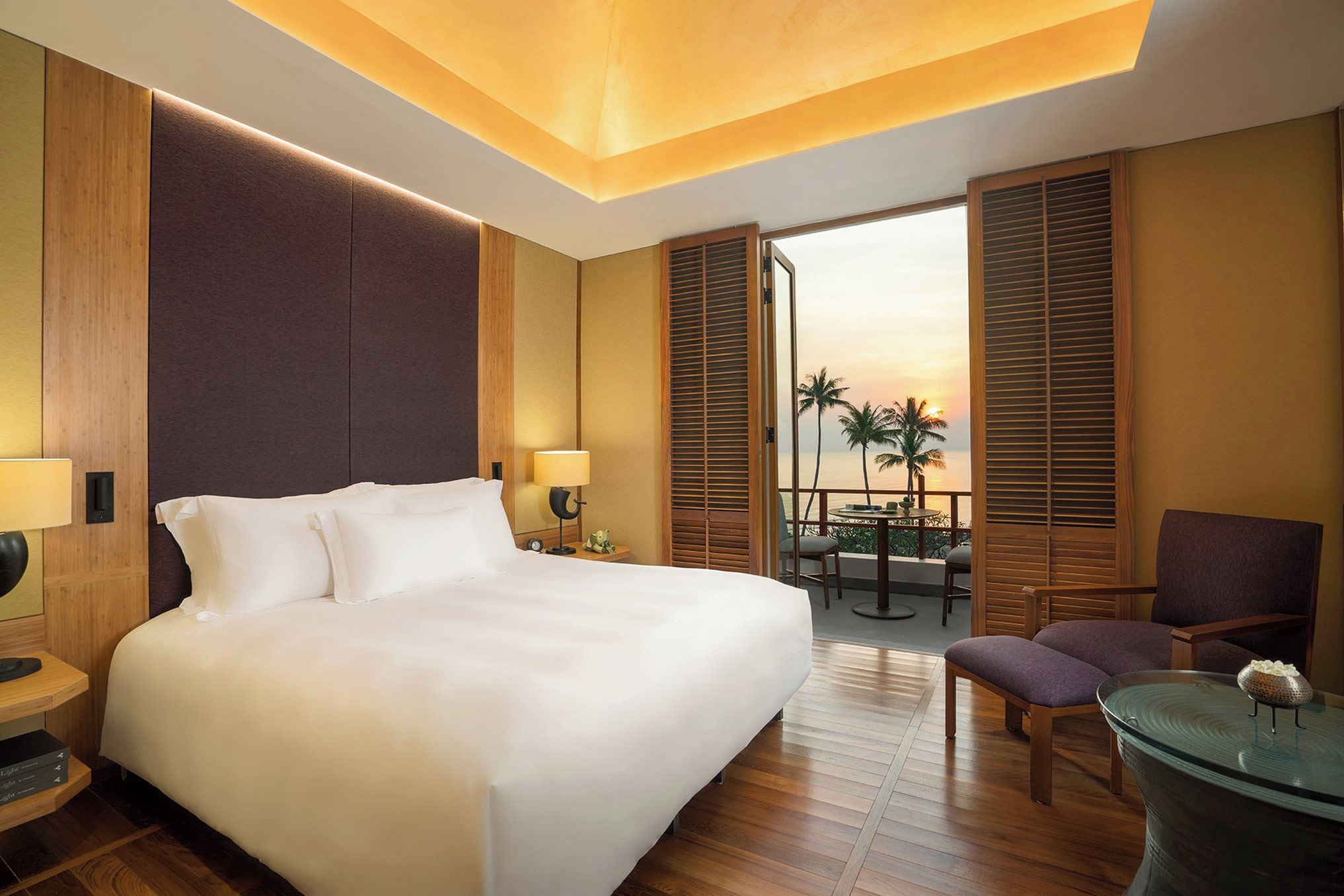 A hotel room with a large bed, a desk, a chair, and a view of palm trees and the sunset.