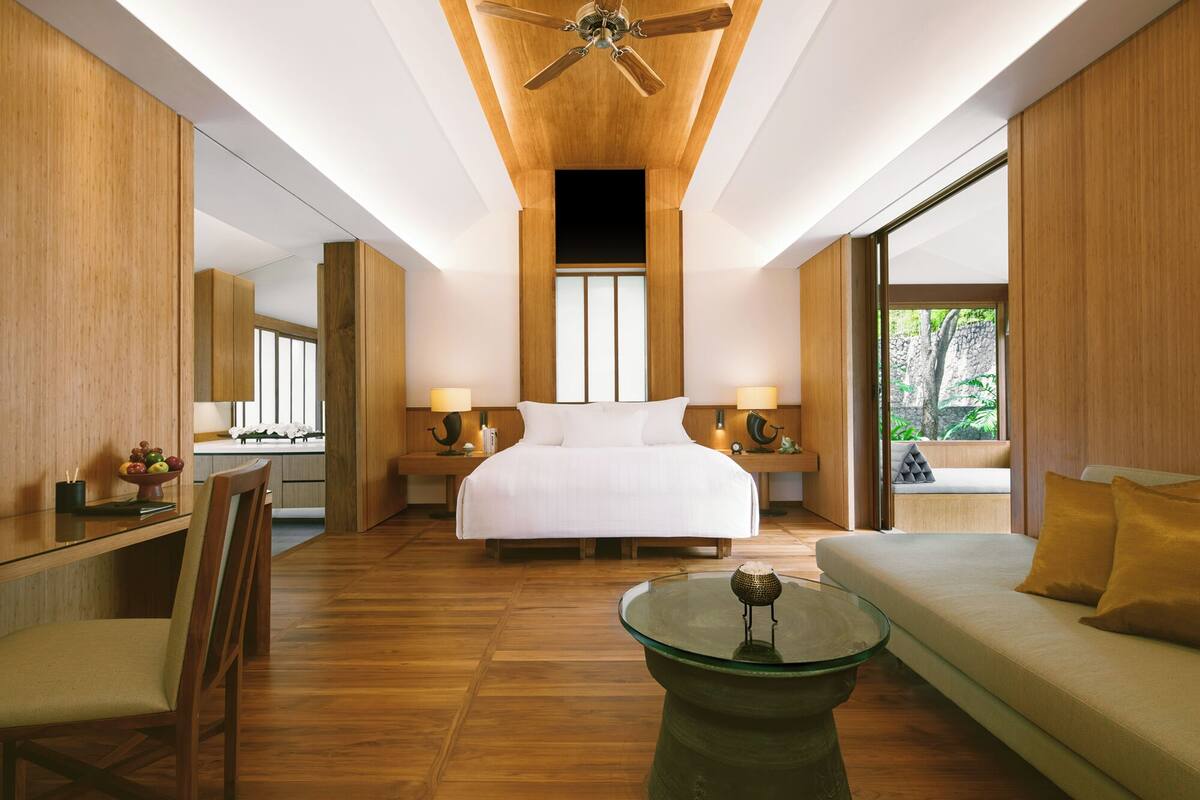 Een moderne hotelkamer met een groot bed, twee nachtkastjes met lampen, een glazen salontafel, een bank met kussens, houten vloeren en een plafondventilator.