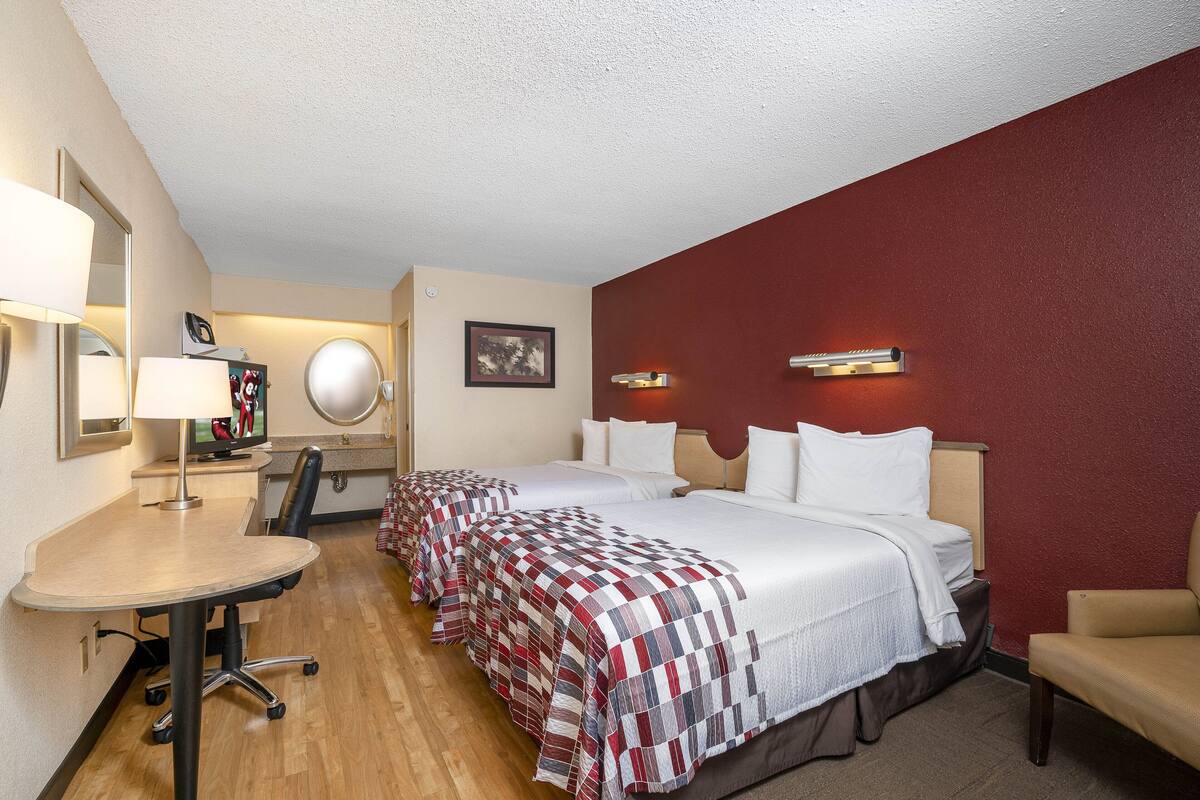 A hotel room with two beds, a desk, a chair, a desk lamp, a TV, and a mirror.