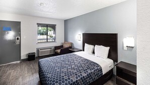 A hotel room with a bed, a chair, a nightstand, a window, and a door. - Rodeway Inn At Fort Lee (Hopewell)