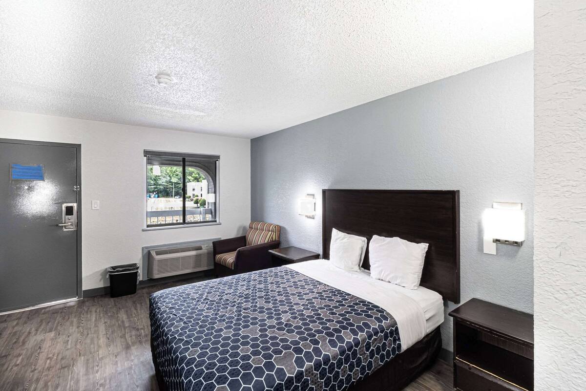 A hotel room with a bed, a chair, a nightstand, a window, and a door.