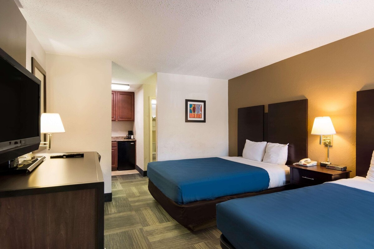 A hotel room with two beds, a desk with a TV, and a small kitchenette.