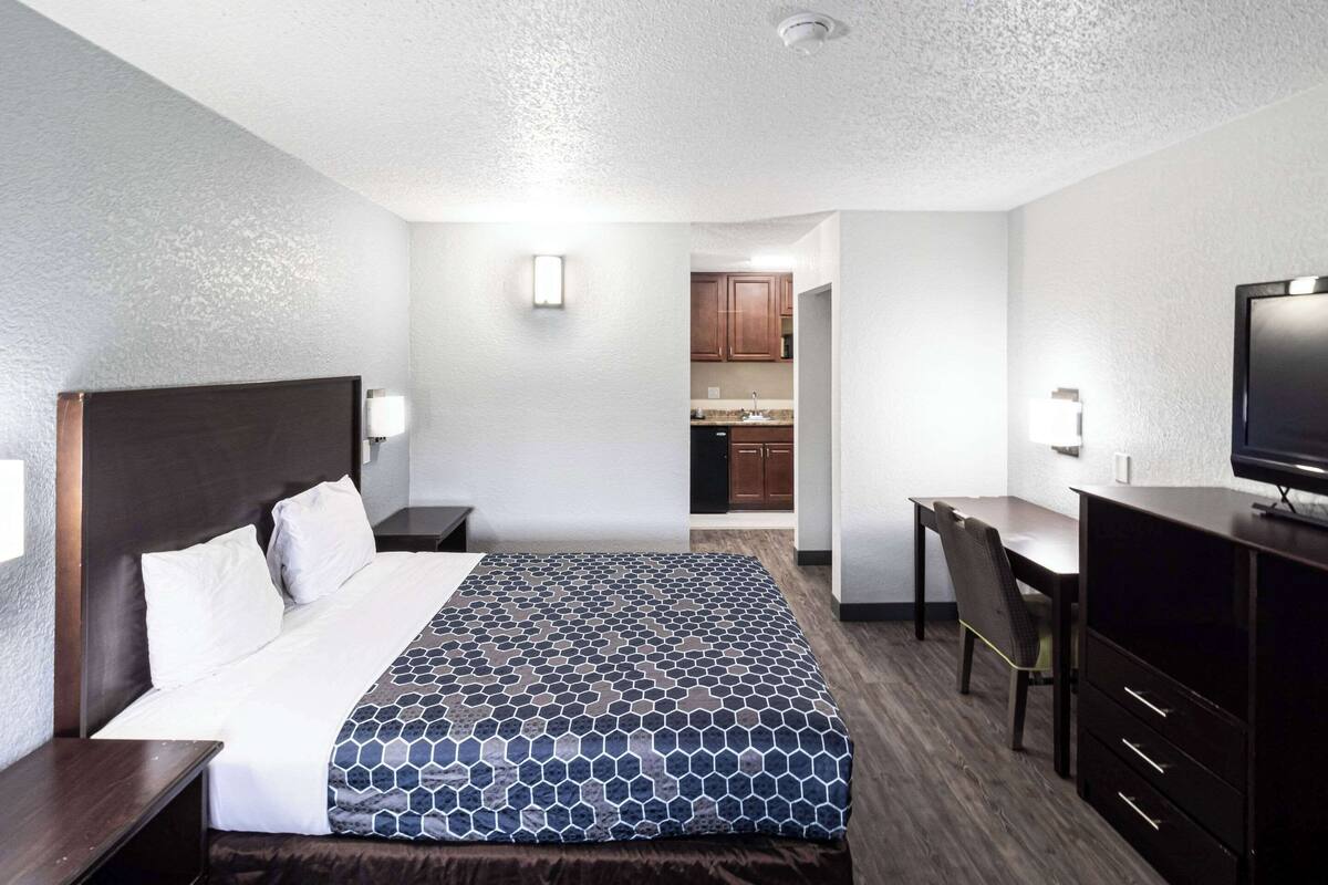 A hotel room with a bed, a desk, a chair, a television, and a kitchenette in the background.