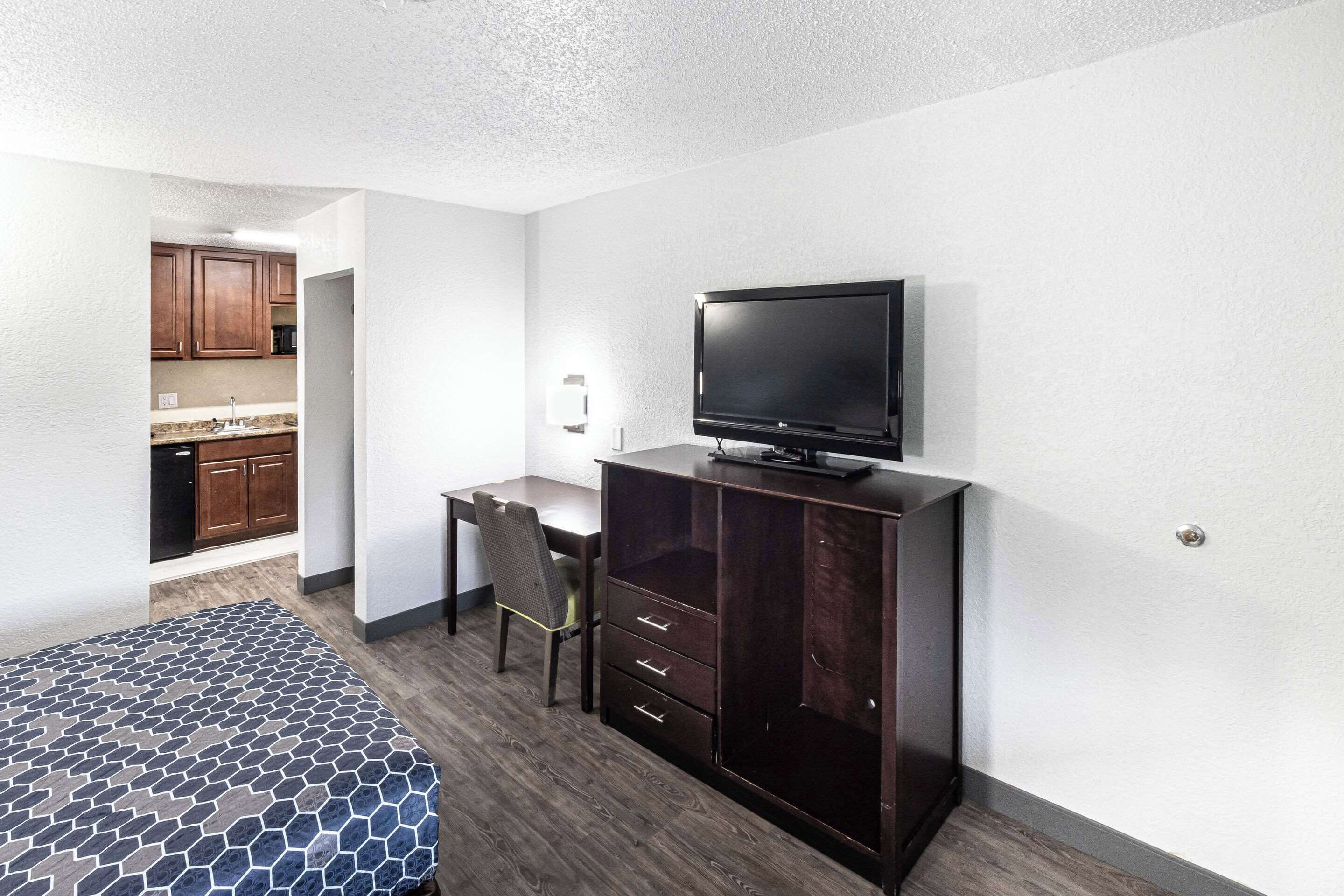 A hotel room with a bed, a desk, a TV, and a wardrobe. There is a kitchenette in the background.