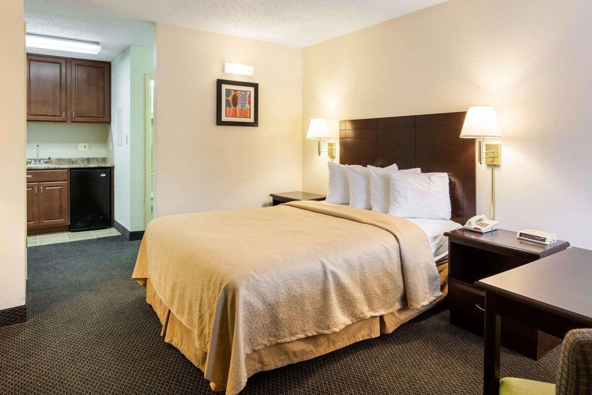 A hotel room with a bed, bedside tables, a desk, a chair, a kitchenette, and a framed picture on the wall.