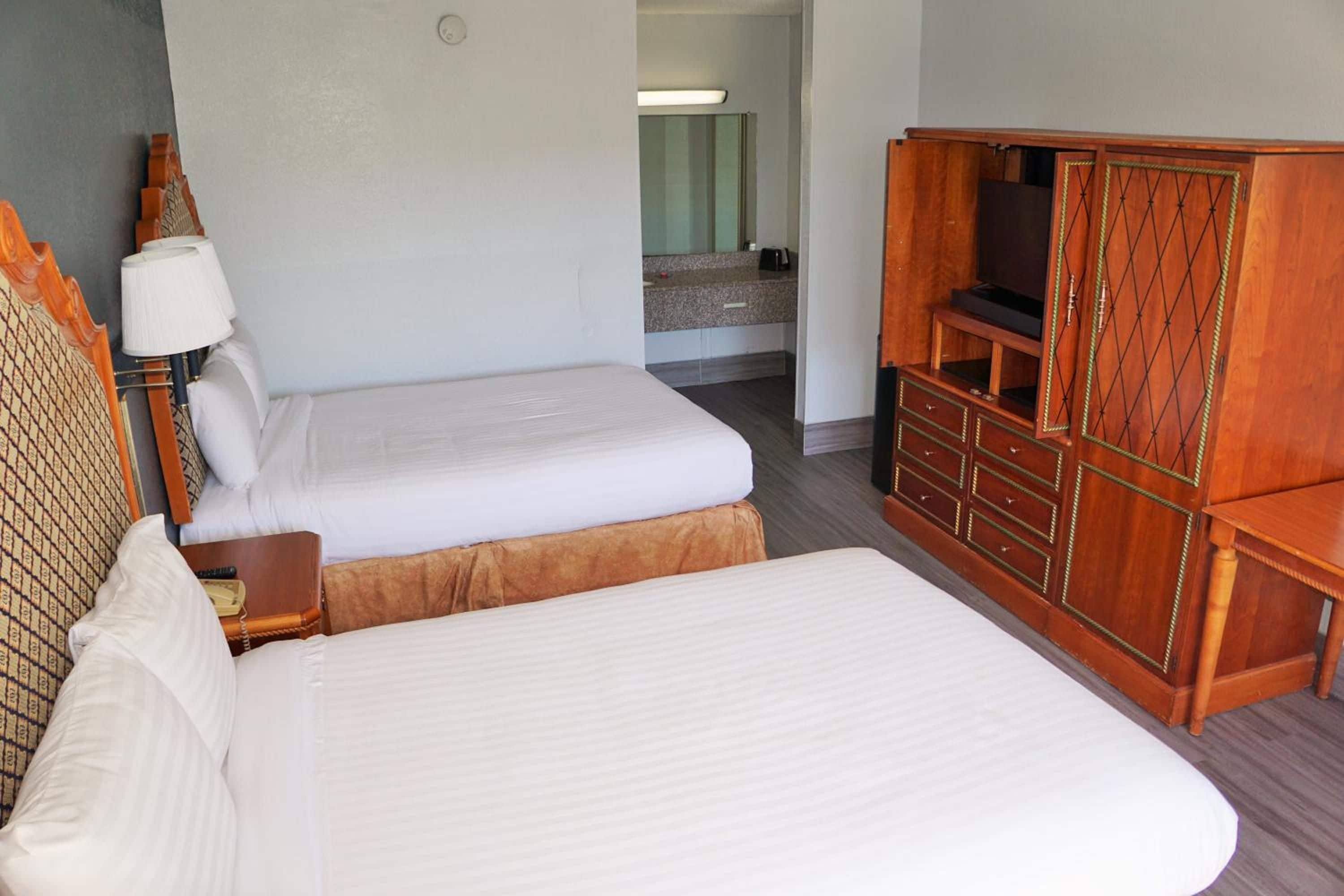 A hotel room with two single beds, a wooden wardrobe, a bedside table with a lamp, and a bathroom visible in the background.