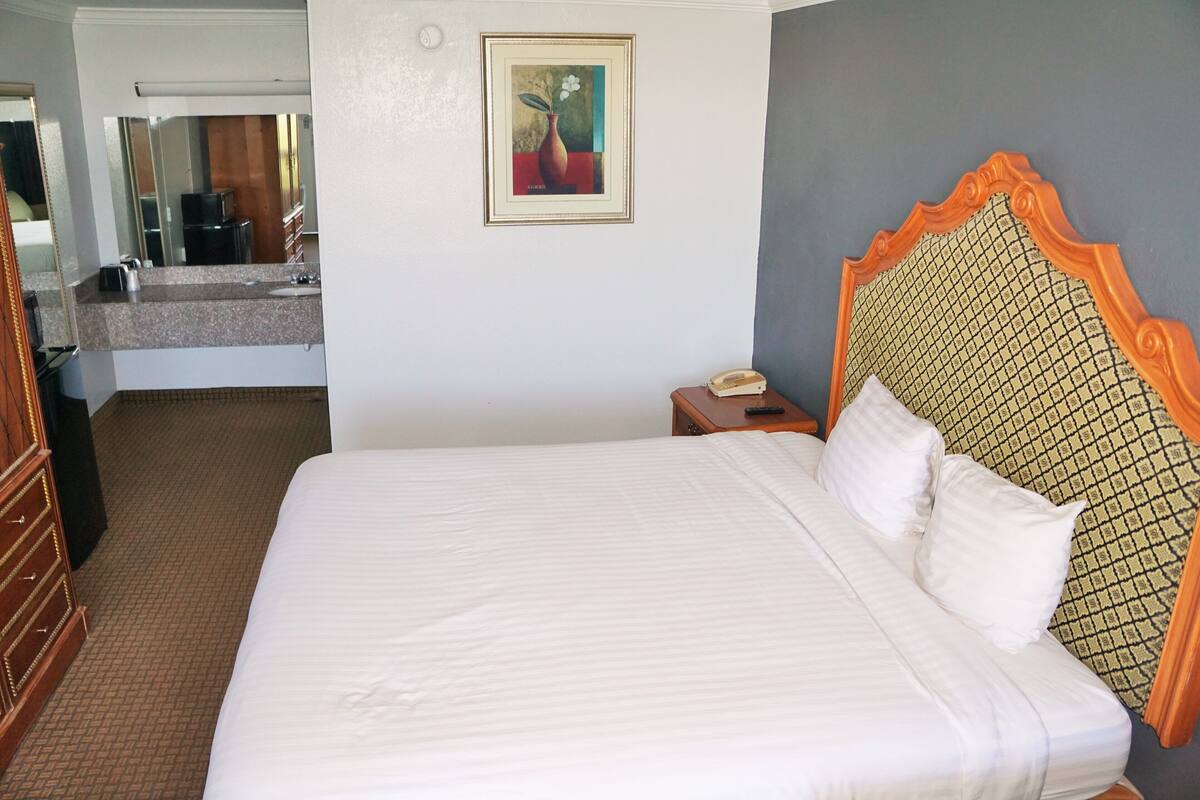 A hotel room with a large bed, a wooden dresser, a nightstand with a telephone, and a painting on the wall.
