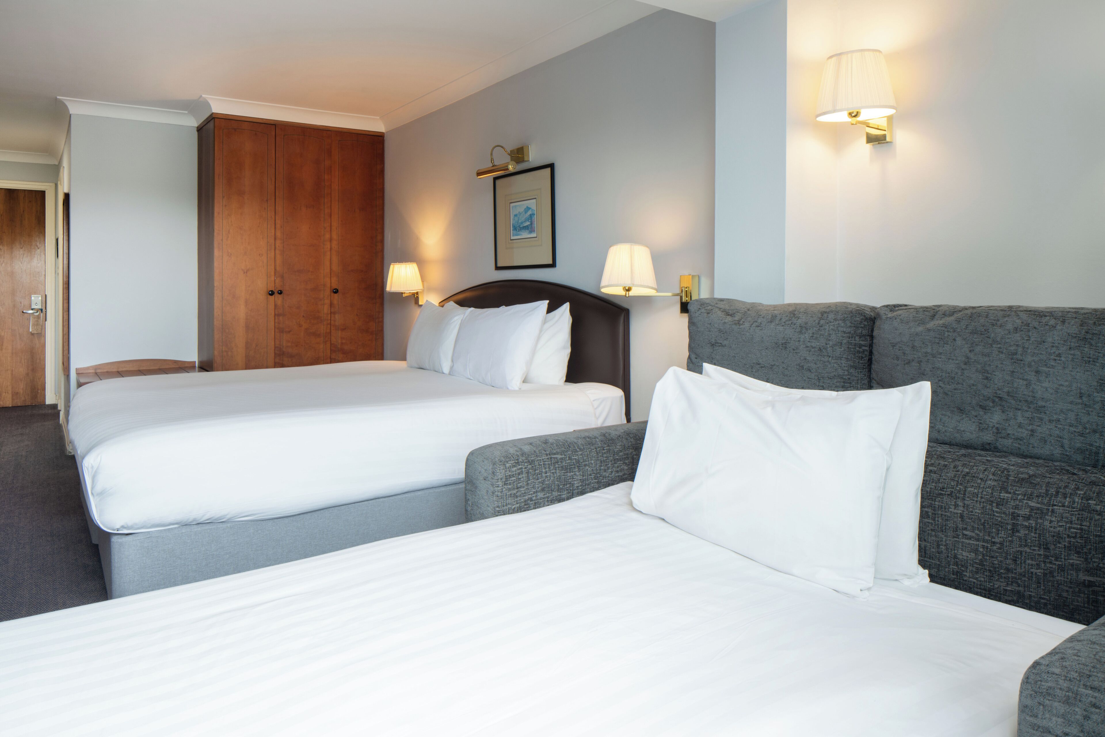 Habitación de hotel con dos camas individuales, un sofá, un armario de madera y dos lámparas de pared.