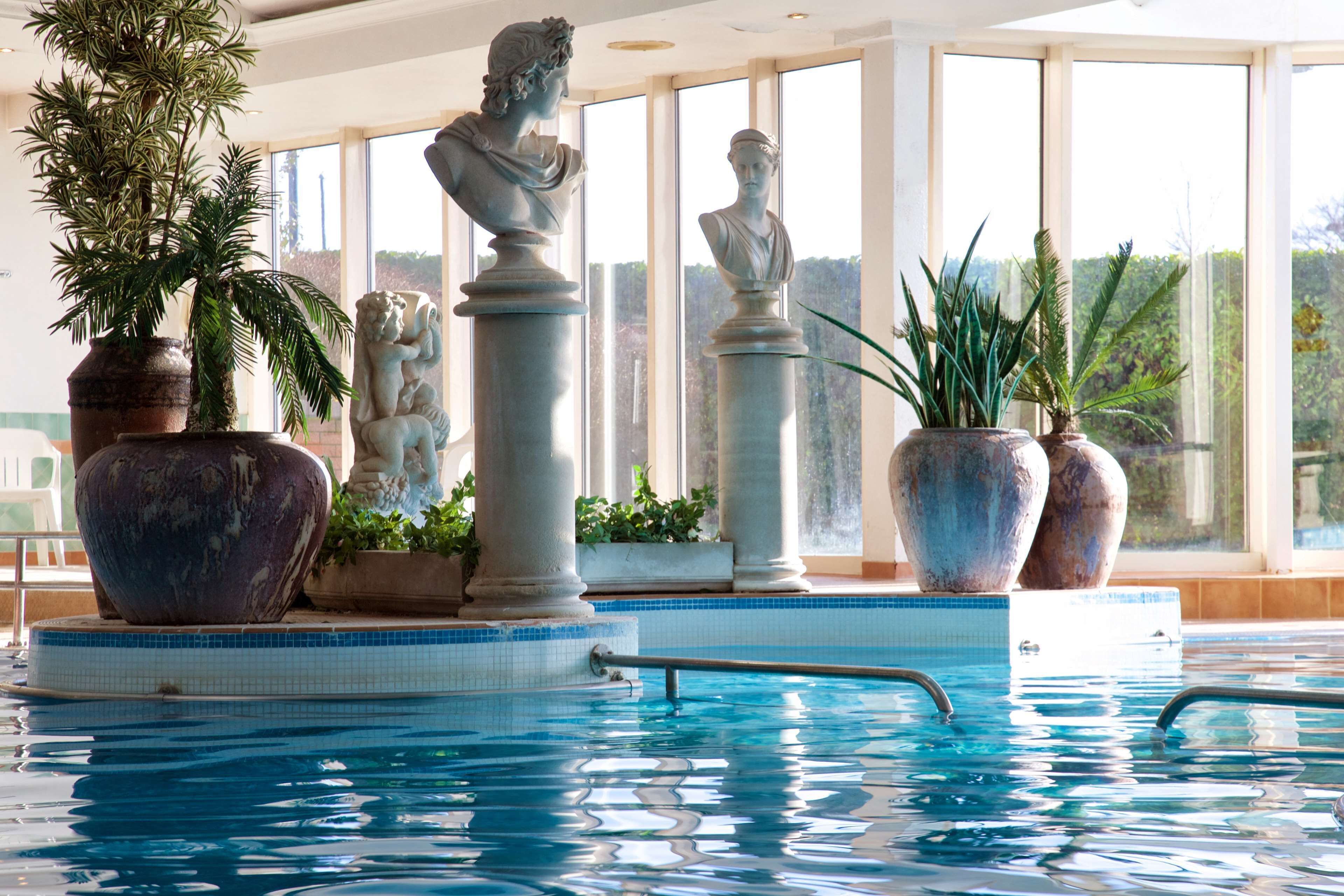 An indoor swimming pool with classical sculptures and large potted plants.