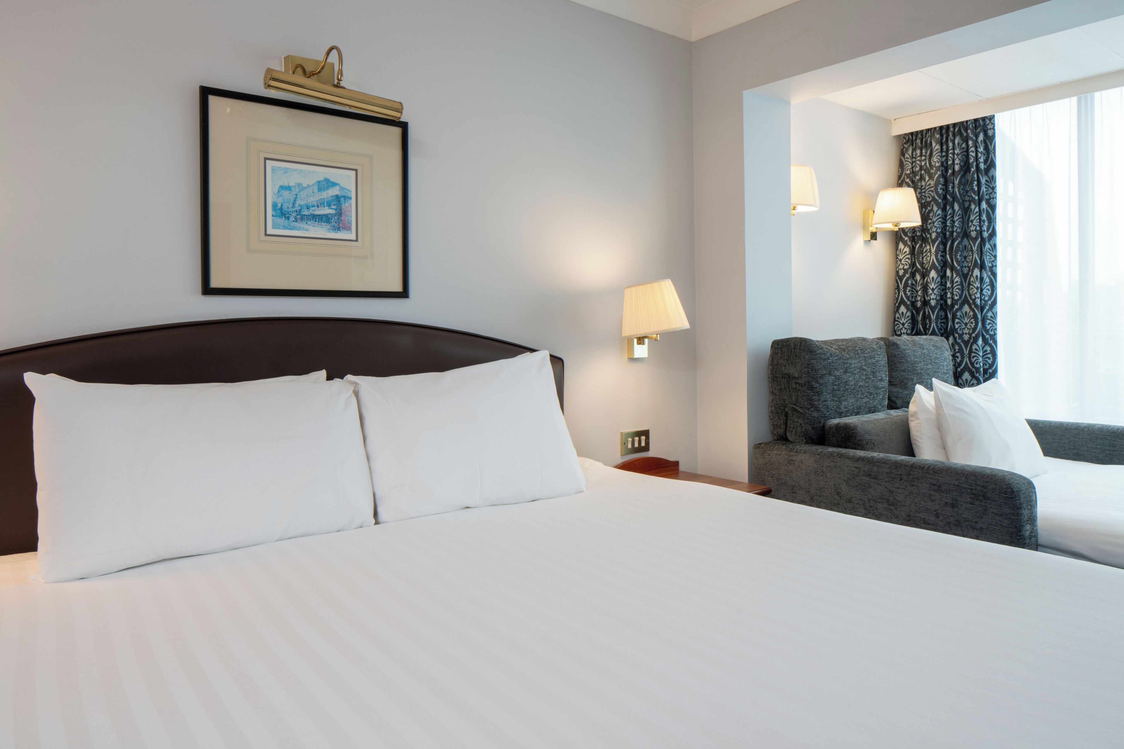 Une chambre d’hôtel avec un grand lit, une chaise, une table de chevet, une lampe, un tableau encadré au mur et une fenêtre avec des rideaux.