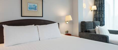 Une chambre d’hôtel avec un grand lit, une chaise, une table de chevet, une lampe, un tableau encadré au mur et une fenêtre avec des rideaux.